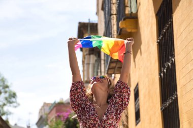 Orta yaşlı sarışın bir kadın, çiçekli bir elbise ve güneş gözlüğü takıyor, sokakta yürüyor, el sallıyor ve eşcinsel gurur bayrağı sallıyor. Kavram lgtbi, gay, lezbiyen, gurur günü.