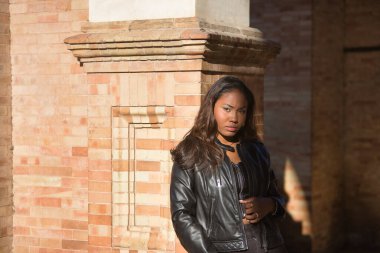 Young, beautiful, Latin and South American woman with top and leather jacket, leaning against a brick wall, posing sensual and attractive between sun and shadow. Concept beauty, fashion, diversity.
