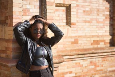 Genç, güzel, Latin ve Güney Amerikalı kadın portresi deri ceketli ve güneş gözlüklü, tuğla duvara yaslanmış, elleri saçlarında, şehvetli ve çekici poz veriyor..
