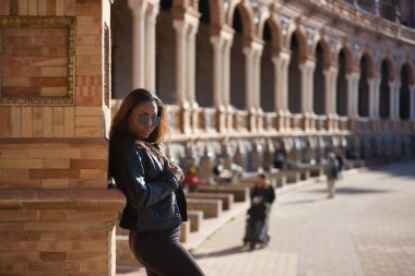Young, beautiful, Latin and South American woman with leather top and jacket, sunglasses and jeans, leaning against a brick wall, posing sensual and attractive. Concept beauty, fashion, diversity.