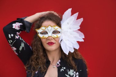 Genç ve güzel bir kadının portresi, sarışın, kıvırcık saçlı ve mavi gözlü, çiçekli ve siyah bluzlu, tüylü beyaz bir karnaval maskeli, kırmızı arka planda.