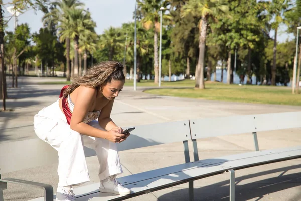아름다운 아메리카 소녀가 벤치에 핸드폰으로 네트워크를 확인하고 있습니다 모바일 스마트폰 — 스톡 사진