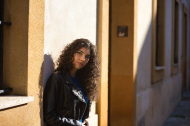 Genç, güzel, esmer, kıvırcık saçlı, siyah deri ceketli ve pantolonlu bir kadın, duvara yaslanmış, güneşin ışınlarını yakalıyor, sakin bir tavırla kameraya bakıyor. Güzellik ve moda kavramı.