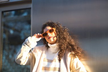 Young woman, beautiful, brunette with curly hair, sweater, coat and sunglasses, leaning on a metal wall, relaxed and calm receiving the sun's rays. Concept beauty, fashion, autumn, winter, cold, relax