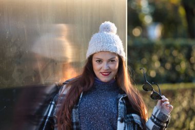 Güzel, düz kahverengi saçlı, süveter, ceket, yün şapka ve güneş gözlüklü, metal bir duvara yaslanmış, gülümseyen ve mutlu bir kadın. Güzellik, moda, sonbahar, kış, soğuk kavramları.