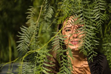 Portrait of beautiful young woman with straight brown hair and brown eyes, looking through small green leaves of a tree. Concept fashion, beauty, trend, plants, looks, leaves, millennial.