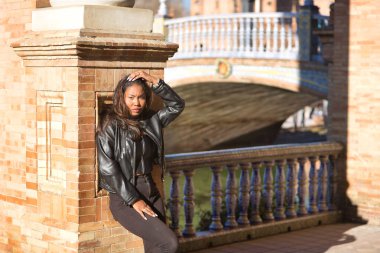 Genç, güzel, Latin ve Güney Amerikalı kadın deri ceketli, bluzlu ve kot pantolonlu, duvara yaslanmış, saçlarına dokunarak kameraya meydan okuyor. Moda kavramı, çeşitlilik.