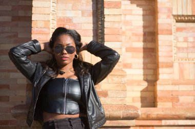 Genç, güzel, Latin ve Güney Amerikalı kadın portresi deri ceketli ve güneş gözlüklü, tuğla duvara yaslanmış, elleri başının arkasında, şehvetli ve çekici poz veriyor..