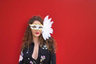 Genç ve güzel bir kadının portresi, sarışın, kıvırcık saçlı ve mavi gözlü, çiçekli ve siyah bluzlu, tüylü beyaz bir karnaval maskeli, kırmızı arka planda.