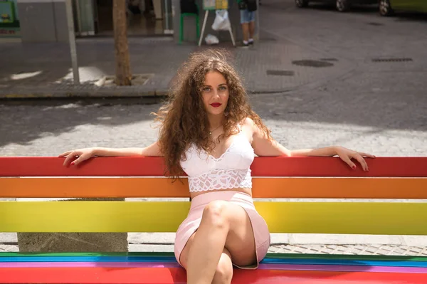 きれいな若い女性は ブロンド 巻き毛と青い目で 白い上とピンクのスカートを着て ベンチにゲイの誇りの色で塗装に座っている コンセプト誇り レズビアン Lgtbi — ストック写真