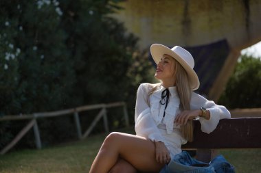 Genç, güzel, yeşil gözlü, beyaz gömlek ve şapka giyen, mini etekli, tahta bir bankta oturan, gülümseyen, rahat ve mutlu bir sarışın kadın. Güzelliği, modayı, trendi, rahatlamayı, mutluluğu kavra.