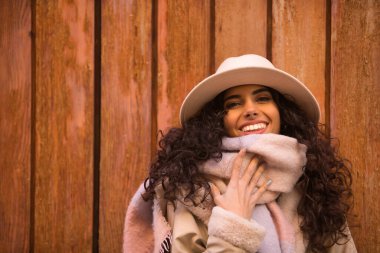 Young, beautiful, brunette woman with curly hair and coat, scarf and hat, looking at camera while protecting herself from cold with scarf, on wooden background. Concept fashion, autumn, winter, cold.