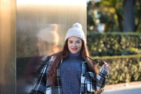 Güzel, düz kahverengi saçlı, kazak, ceket, yün şapka ve güneş gözlüklü, metal bir duvara yaslanmış, cilveli ve şehvetli bir kadın. Güzellik, moda, sonbahar, kış, soğuk kavramları.