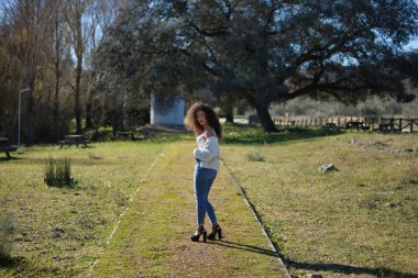 Genç, güzel, esmer, kıvırcık saçlı, beyaz bluzlu, kot ve topuklu giyen, parkın ortasında kameraya bakarak poz veren bir kadın. Güzelliği, modayı, pozu kavra.