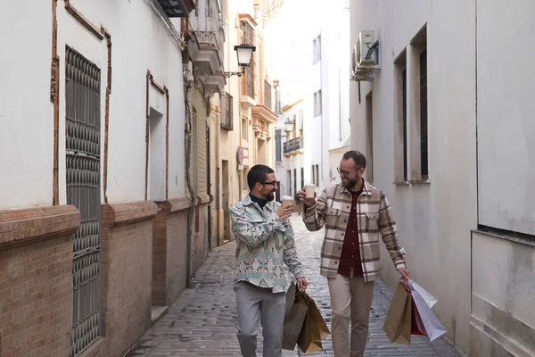 stock image Real marriage of gay couple, walking along a lonely street, with a glass of coffee in one hand and the other full of shopping bags. Concept lgtb, lgtbiq+, couples, in love, shopping, sales.