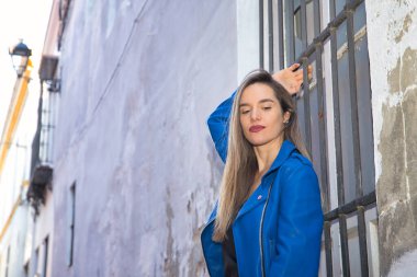 Young woman, beautiful and blonde, wearing a black shirt and blue leather jacket clutching the grille of a window, smiling sensually and carefree. Concept sensuality, provocation.