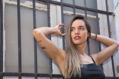 Genç bir kadın, güzel ve sarışın, siyah kolsuz tişörtlü, pencerenin ızgarasına yapışmış, şehvetli ve çapkın bakışlı. Duygusallık, güzellik, flört..