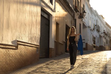Young woman, beautiful and blonde, wearing a black tank top, blue fur jacket in her hand and jeans, walking down a lonely and lost street during the golden hour. Concept fashion, beauty, sunset.