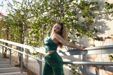 Latin ve İspanyol kız, genç ve asi, yeşil kadife bluzlu ve pantolonlu, şehvetli ve kışkırtıcı bir tavırla poz veriyor. Güzelliği, modayı, şehveti, provokasyonu kavra.