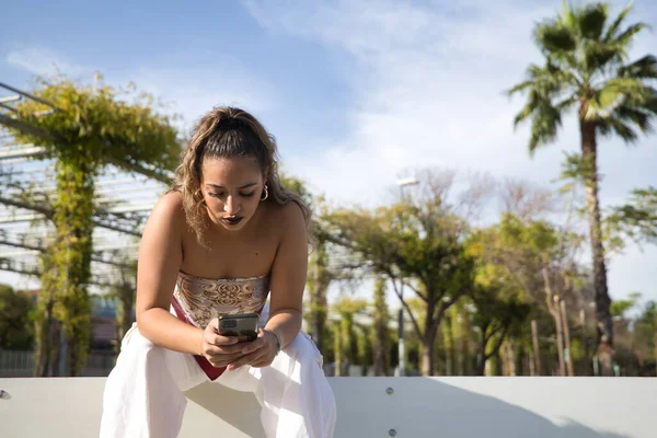 ラテン系とヒスパニック系の少女 若く美しい 彼女の携帯電話上のソーシャルネットワークをチェック ベンチに座って モバイルコンセプト スマートフォン コンサルティング アプリ — ストック写真