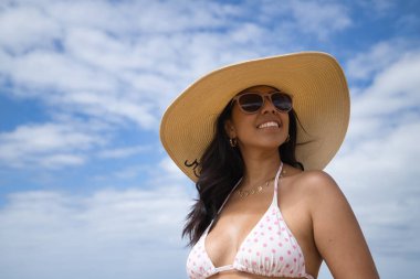 South American woman, young and beautiful, brunette with sunglasses, hat and bikini posing happy and smiling on the beach. Concept sea, sand, sun, beach, vacation, travel, summer.