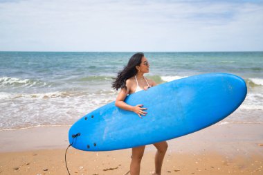 South American woman, young and beautiful, brunette with sunglasses and bikini, posing while holding a blue surfboard. Concept sea, sand, sun, beach, vacation, surf, summer.
