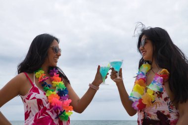 Two young, beautiful, brunette South American women in bikinis, sunglasses and flower necklaces toasting with glasses of blue wine. Concept vacation, friends, summer, beach, drinks.