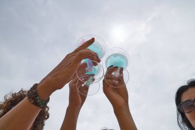 Aşağıdan görünen sahilde kadeh kaldıran üç kadının tuttuğu üç mavi şarap kadehinin ayrıntıları. Tatil, yaz, plaj, içkiler.