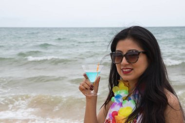 Genç ve güzel Güney Amerikalı kadın, esmer, güneş gözlüğü, bikini ve çiçek kolyesi elinde bir bardak mavi şarapla sahilde. Yaz konsepti, plaj, içkiler.