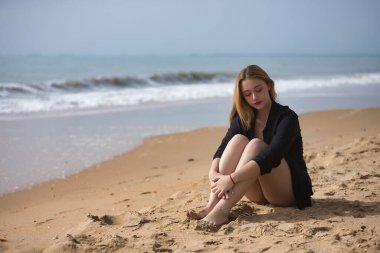 Genç, sarışın, bikinili ve siyah tişörtlü güzel bir kadın sahilde oturuyor, üzgün, yalnız, üzgünüm, depresyonda. Yalnızlık, üzüntü, keder, depresyon kavramını.