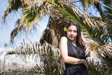 Genç ve çekici bir kadın, mavi gözlü, siyah mayosu ve elbisesi olan, şehvetli ve kışkırtıcı bir tavırla poz veren, arkasında palmiye ağaçları olan. Duygusallık, provokasyon, plaj.