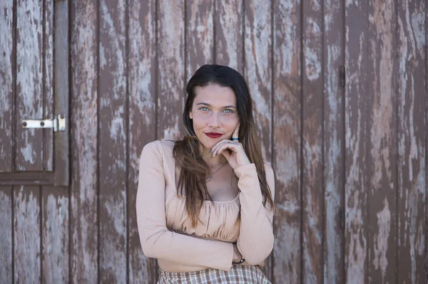 stock image Attractive young woman, with blue eyes, sweater and plaid skirt, with her hand on her chin in a pensive attitude, on a wall of wooden planks. Concept thoughts, expressions.