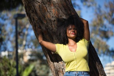 Genç bir kadın, güzel ve siyah saçlı, sarı gömlekli ve kotlu, şehvetli ve kışkırtıcı bir tavırla bir ağacın gövdesine yaslanmış. Duygusallığı, provokasyonu, flörtü kabul et..