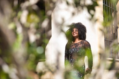 Genç bir kadın, güzel ve siyah saçlı, afro saçlı, çiçekli elbisesi duvara dayanmış, gülümseyen ve mutlu, yeşil bir bitkinin içinden görülüyor. Güzelliği kavra, kahkahayı, mutluluğu, eğlenceyi, bitkileri.