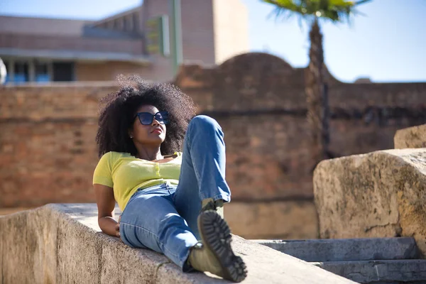 美しい アフロの髪を持つ黒の女性は 黄色のTシャツ ジーンズやサングラスを着て 石の壁に横たわって リラックスした 穏やかな日光浴 休暇の概念 — ストック写真