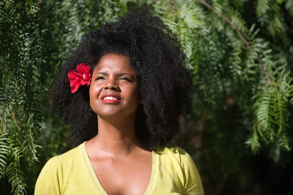 Afro saçlı, sarı tişörtlü ve yeşil bitkilerin arka planında kırmızı çiçekli genç, güzel, siyah bir kadının portresi. Güzellik, seyahat, tropikal, bitkiler ve çiçekler.