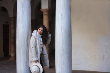 Genç, güzel, kıvırcık saçlı esmer kadın, bej kazak, ceket ve şapka, mermer sütunlara yaslanmış, saçlarına dokunuyor, sakin ve rahat. Güzellik, moda, sonbahar, kış, soğuk, rahatla.