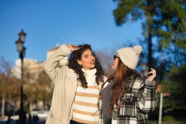 Two women, young and beautiful, with sweater, sunglasses, coat and one with wool hat, embraced, looking at each other, very smiling and happy. Concept autumn, winter, cold, friendship, full happiness.