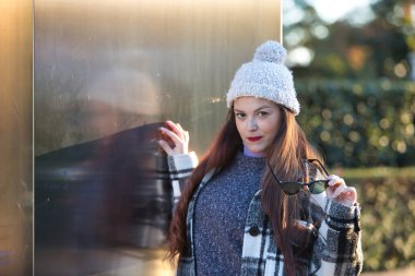 Güzel, düz kahverengi saçlı, kazak, ceket, yün şapka ve güneş gözlüklü, metal bir duvara yaslanmış, cilveli ve şehvetli bir kadın. Güzellik, moda, sonbahar, kış, soğuk kavramları.