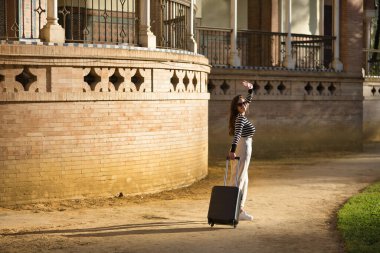 Genç ve güzel bir kadın, İspanyol, kahverengi saçlı, çizgili kazaklı, keten pantolonlu ve güneş gözlüklü, gün batımında valizinin yanında, arkasından el sallayarak veda ediyor. Seyahat, tatil, bavul