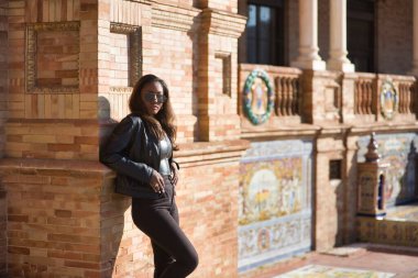 Young, beautiful, Latin and South American woman with leather top and jacket, sunglasses and jeans, leaning against a brick wall, posing sensual and attractive. Concept beauty, fashion, diversity.