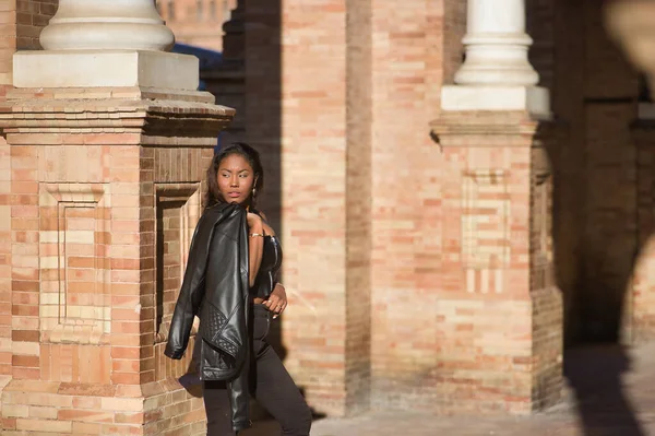 stock image Young, beautiful, Latin and South American woman in leather top and jeans, with jacket over her shoulder, leaning against a brick wall, posing sensual and attractive. Concept fashion, diversity.