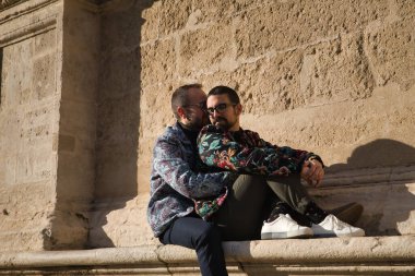 Biri diğerini çok sevecen, suç ortağı ve mutlu bir tavırla kucaklarken, taştan bir duvarda oturan eşcinsel çiftin gerçek evliliği. Kavram lgtb, lgtbiq +, çiftler, aşık, aşık.