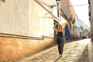 Young woman, beautiful and blonde, wearing a black tank top, blue fur jacket in her hand and jeans, walking down a lonely and lost street during the golden hour. Concept fashion, beauty, sunset.