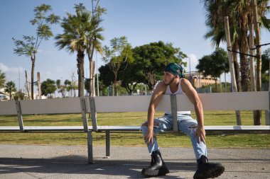 Latin ve İspanyol çocuk, genç, asi, başörtüsü ve askeri botları olan, kaybolmuş görünüşlü, üzgün, yalnız, bankta oturan. Sorunlu konsept, asi, çete, rap, yalnızlık.