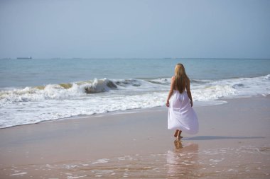 Genç, güzel, beyaz elbiseli sarışın bir kadın, yalnız bir sahilde yürüyor, rahat ve sakin, arkadan görülüyor. Huzuru, huzuru, yalnızlığı, kendini bulmayı kabul et..