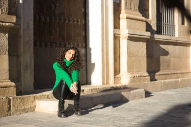 Genç, güzel, kıvırcık saçlı, yeşil boğazlı kazaklı, deri pantolonlu, siyah çizmeli, sakin ve rahat bir şekilde merdivende oturan esmer bir kadın. Güzellik, moda, sonbahar, kış, soğuk kavramları.