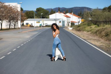 Genç, güzel, kıvırcık saçlı, beyaz tişörtlü, kot pantolonlu ve topuklu ayakkabılı, yalnız bir yolun ortasında poz veren esmer bir kadın. Güzellik, moda, poz, model kavramı.
