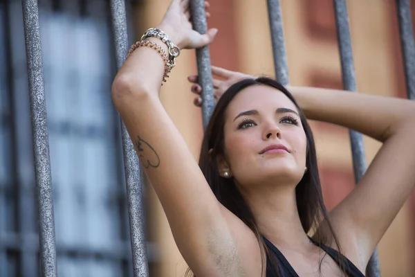 stock image Young and beautiful woman with straight brown hair, wearing an elegant black dress, clinging to the bars of a fence with a distracted look. Concept fashion, beauty, distraction, thoughts.