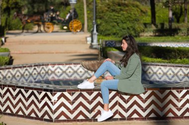 Güzel bir genç kadın, İspanyol, kahverengi saçlı, yeşil ceket ve kot pantolon giyiyor, yalnız, rahat ve sakin, çeşmede oturuyor. Yalnızlık kavramı, rahatlama, sakinlik..